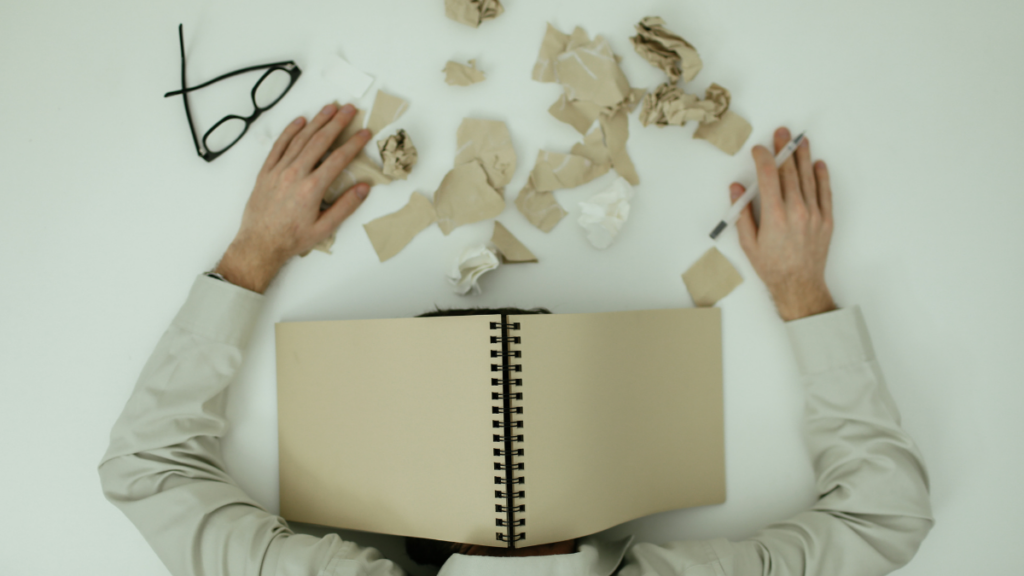 Person resting their head beneath an open notebook at a desk scattered with crumpled paper.